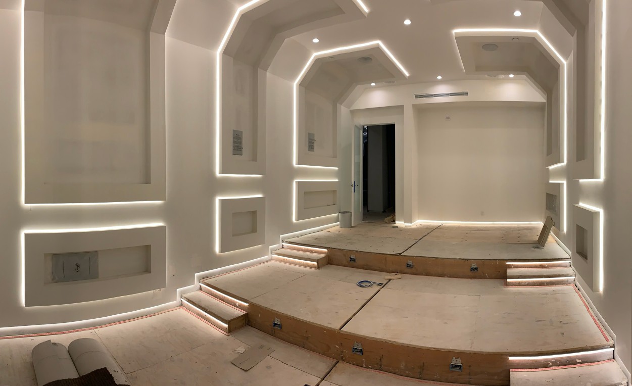 Theater room mid-construction with linear coves energized over plywood subfloor and finished drywall.